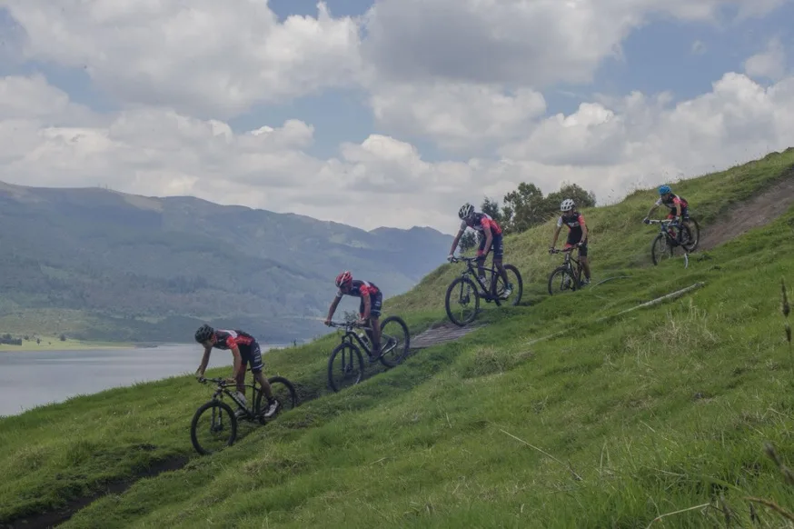 Cinco ciclistas de ONE descendiendo en fila una ladera de pasto sobre el embalse