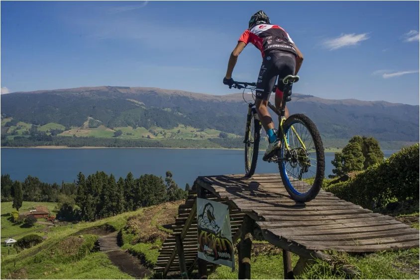 Ciclista de ONE de pie sobre los pedales, con el sillín libre entre las piernas, encarando una rampa de madera sobre el embalse