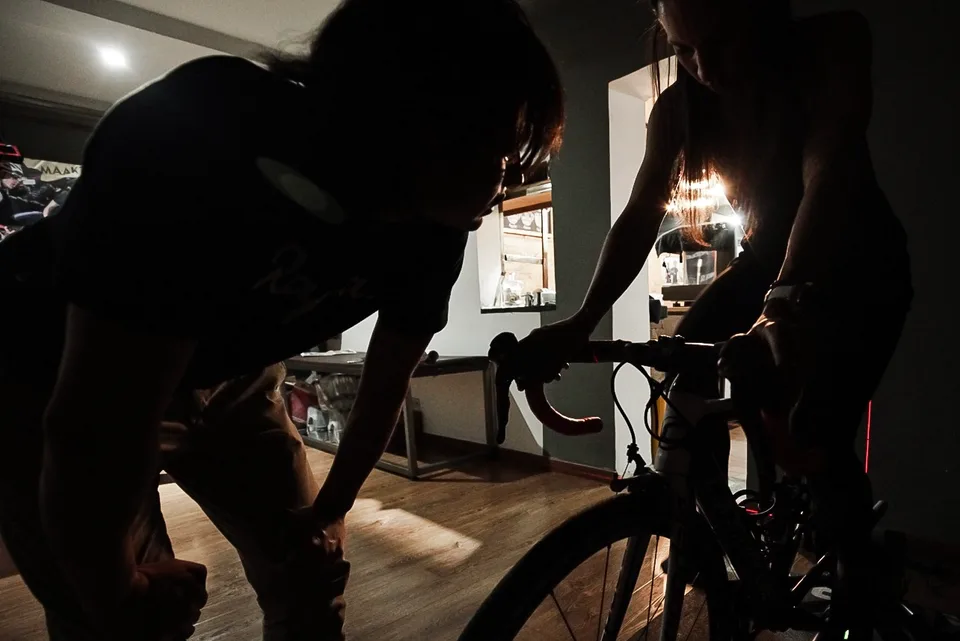 Ciclista pedaleando en rodillo en una habitación a media luz mientras el entrenador observa el esfuerzo